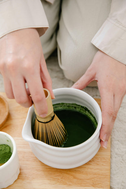 Person preparing matcha green tea using a whisk in a white bowl on a wooden surface.