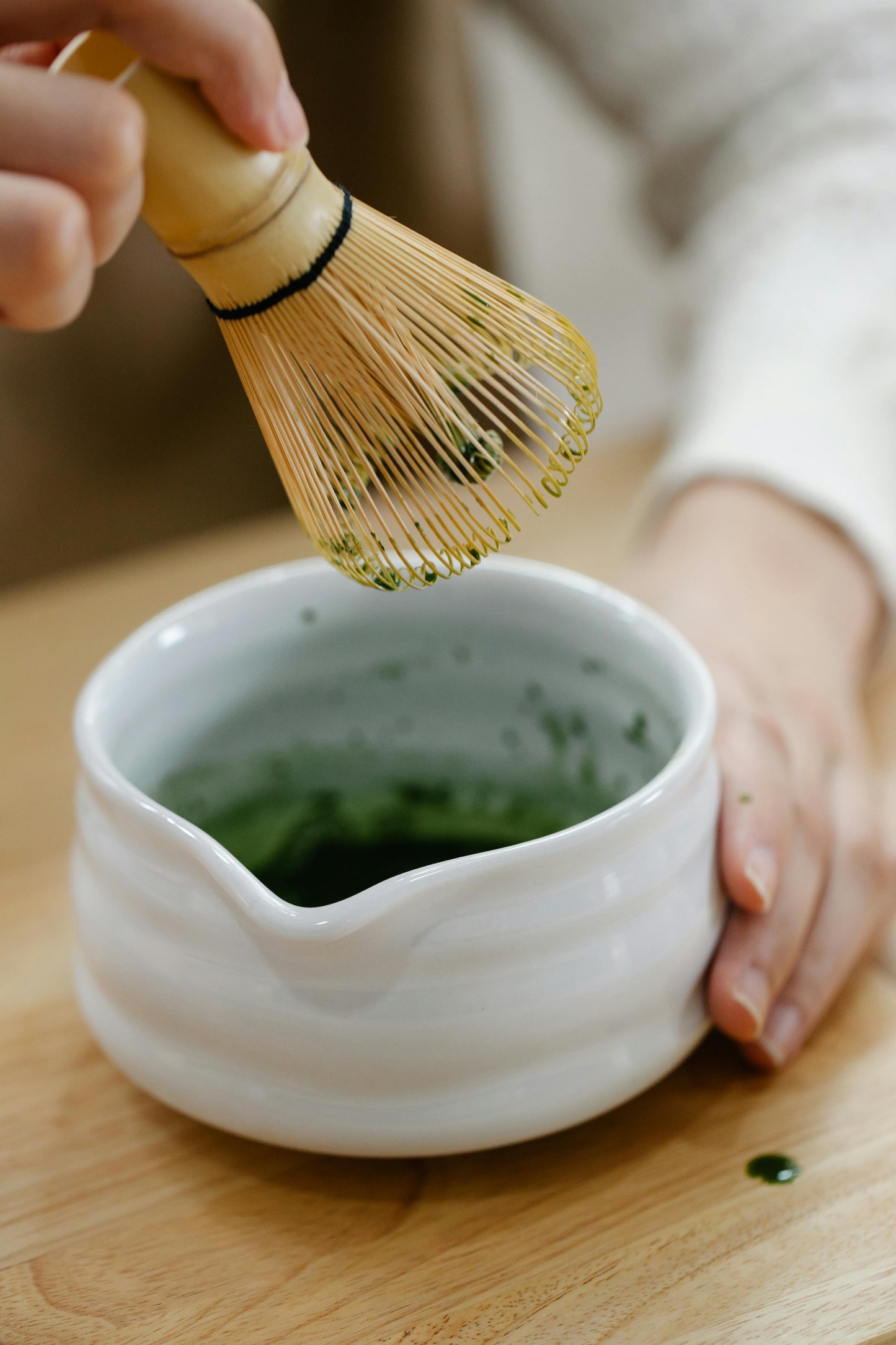 Person using a bamboo whisk to prepare matcha green tea in a white ceramic bowl on a wooden surface.