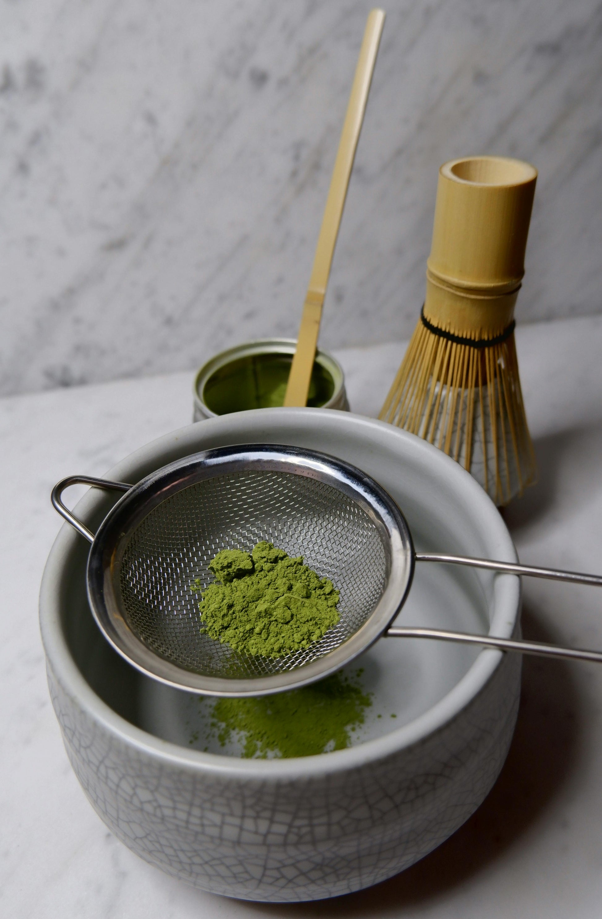 Matcha green tea preparation with a bowl, whisk, and sieve on a marble surface.